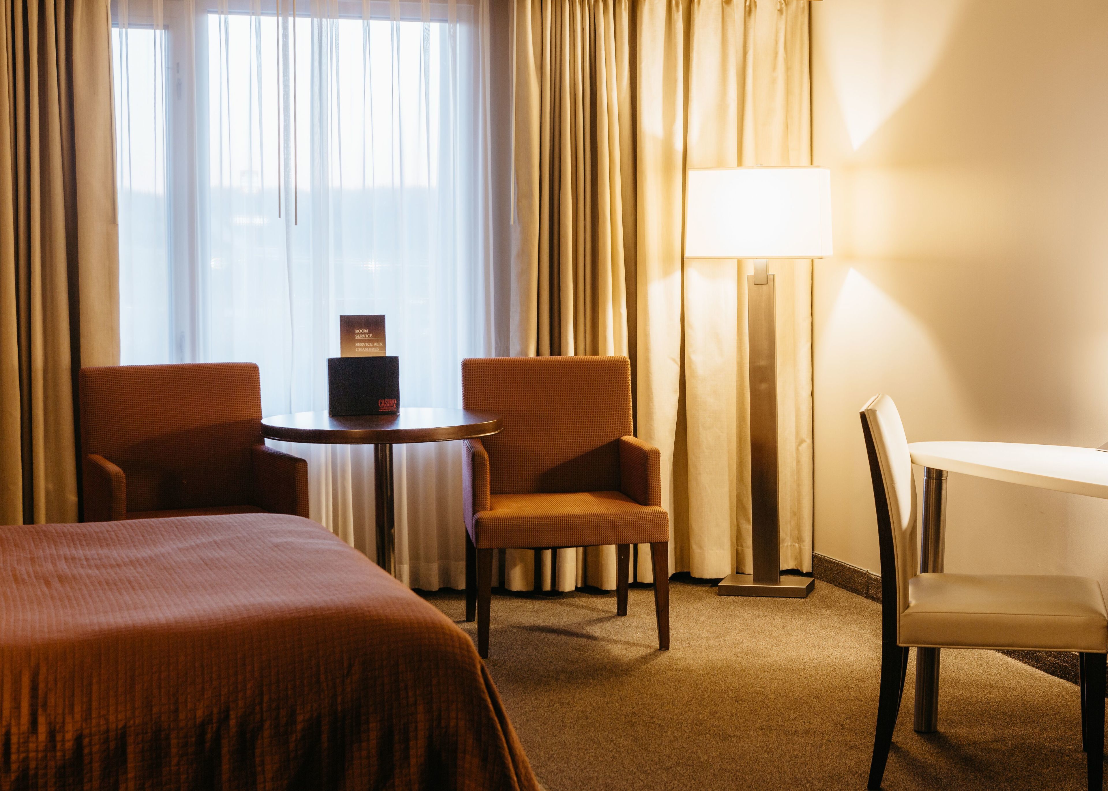 A hotel room with a bed, two armchairs, a small table, a desk, a lamp, and a sign indicating room service availability.