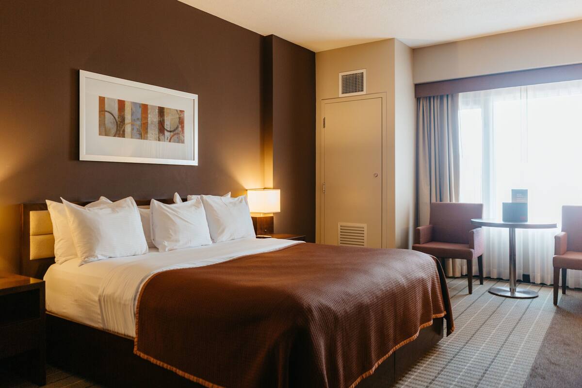 A hotel room with a large bed, two bedside tables, a framed artwork on the wall, a wardrobe, a seating area with a table and chairs, and a window with curtains.