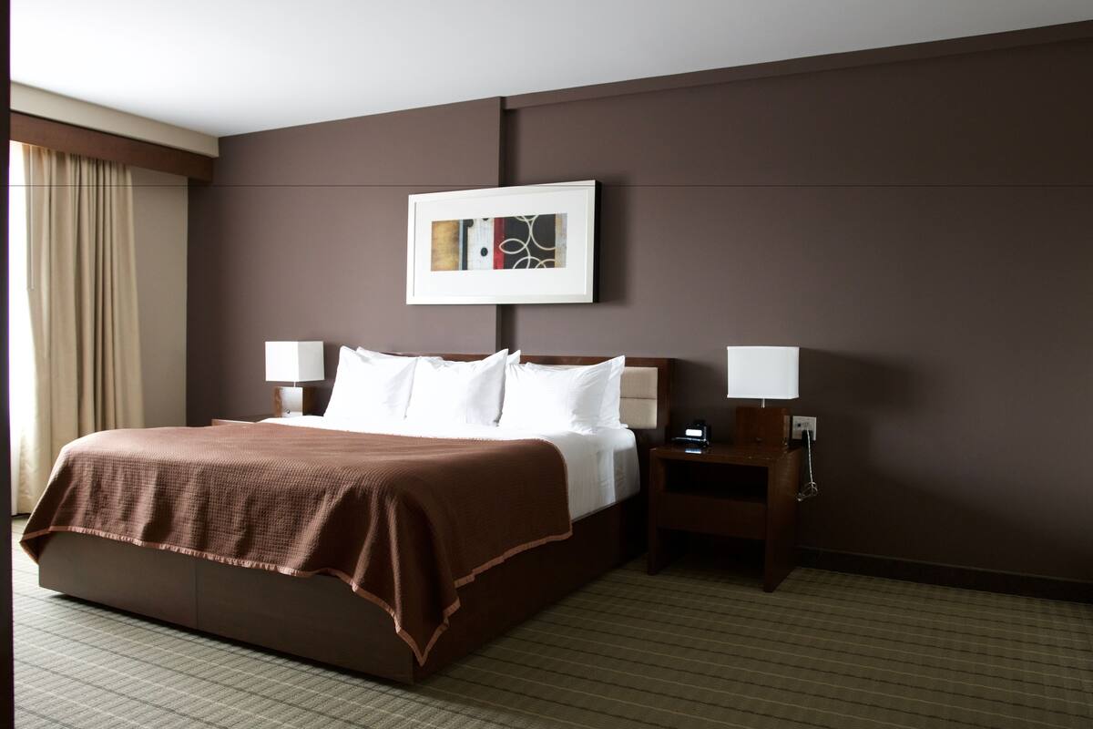 A hotel room with a large bed, two bedside tables, a nightstand, a framed picture on the wall, and a window with curtains.