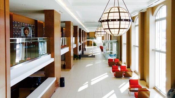 A spacious hotel lobby with a long corridor, red seating areas, large windows, and modern lighting fixtures.