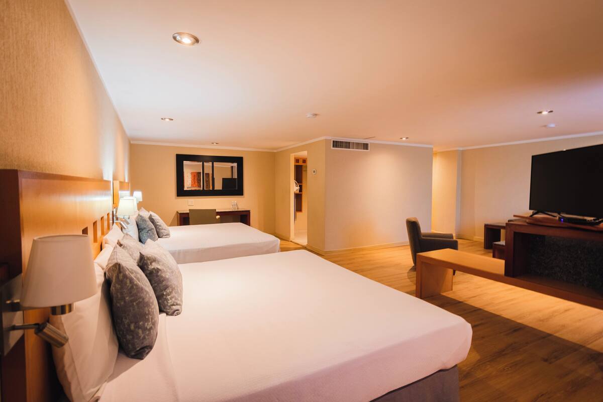 A hotel room with two beds, a desk, a chair, a television, and a bench.
