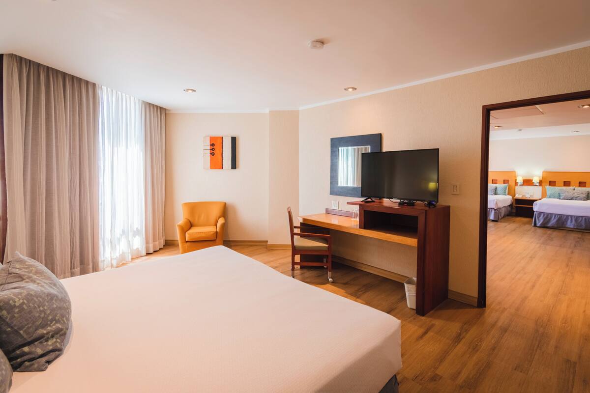 A hotel room with a large bed, a desk with a TV, a chair, and a view of another room.