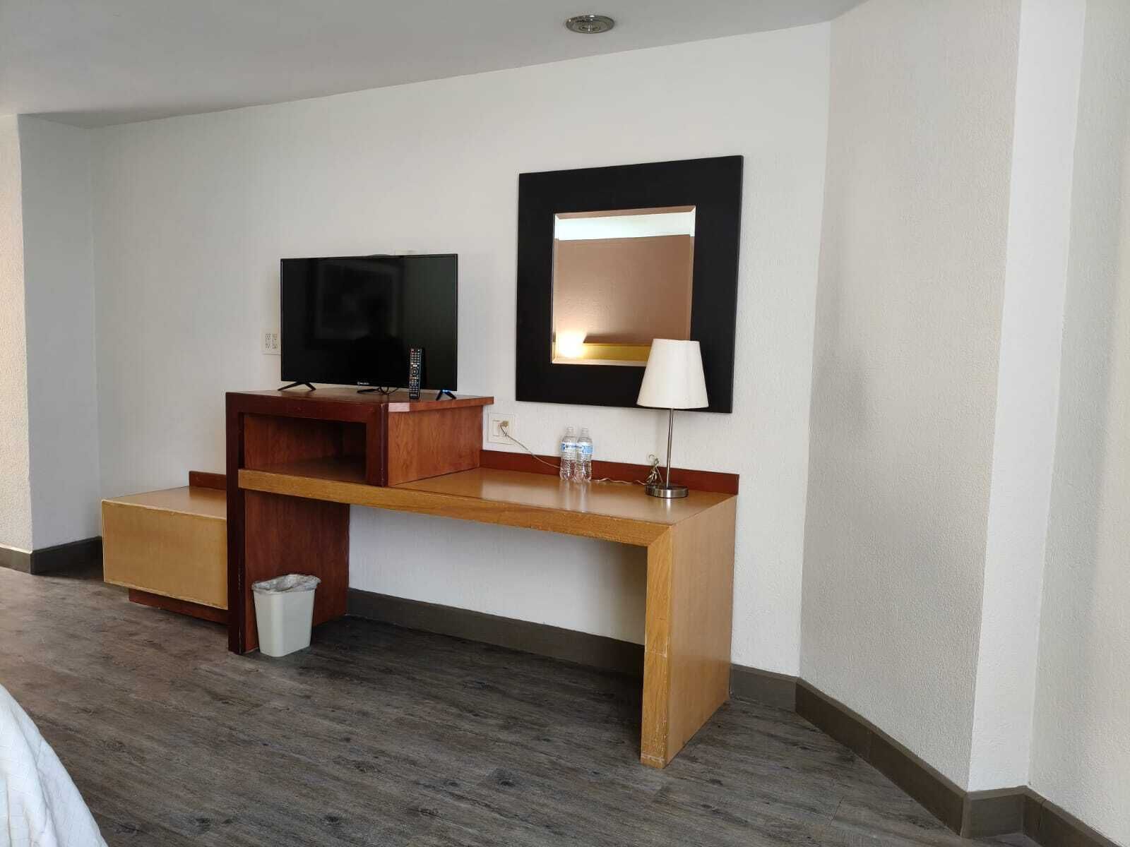 A hotel room with a wooden desk, a television, a mirror, and a lamp.