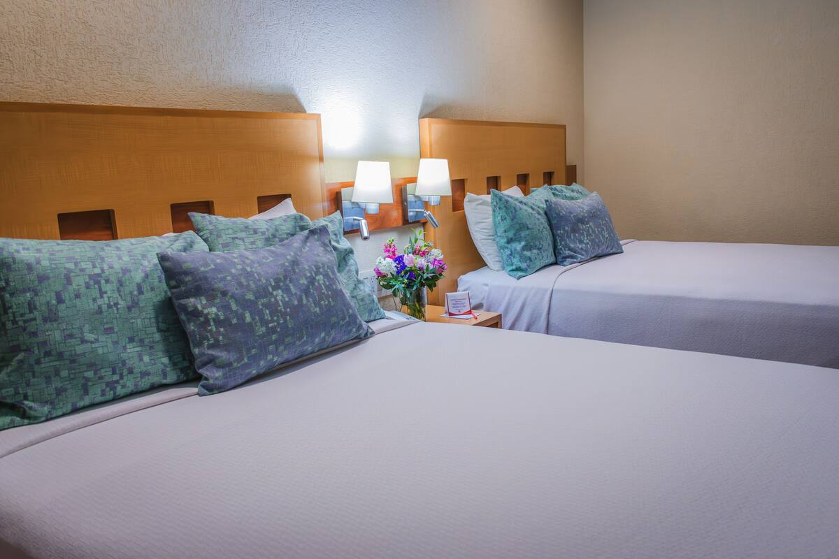 A hotel room with two beds, each with a headboard, pillows, a nightstand with a lamp, and a vase with flowers.