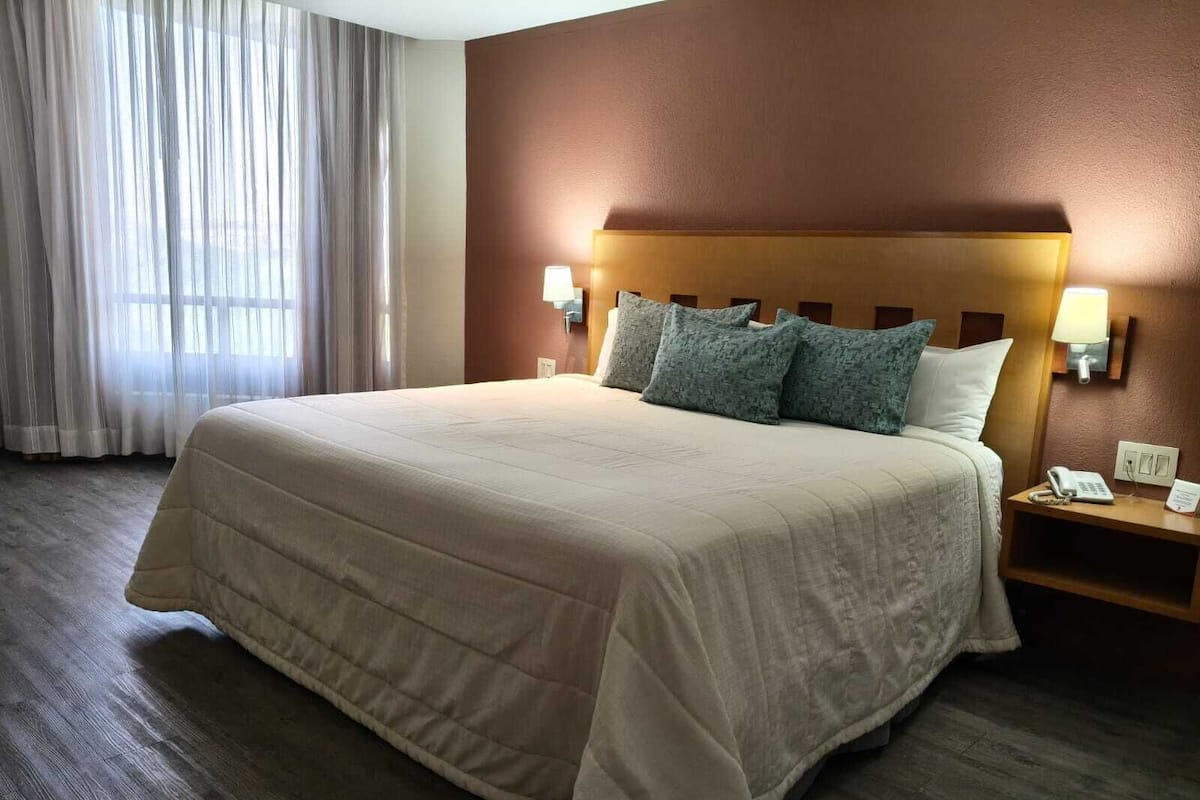 A hotel room with a large bed, wooden headboard, two bedside lamps, a nightstand with a phone and a sign, and a window with sheer curtains.