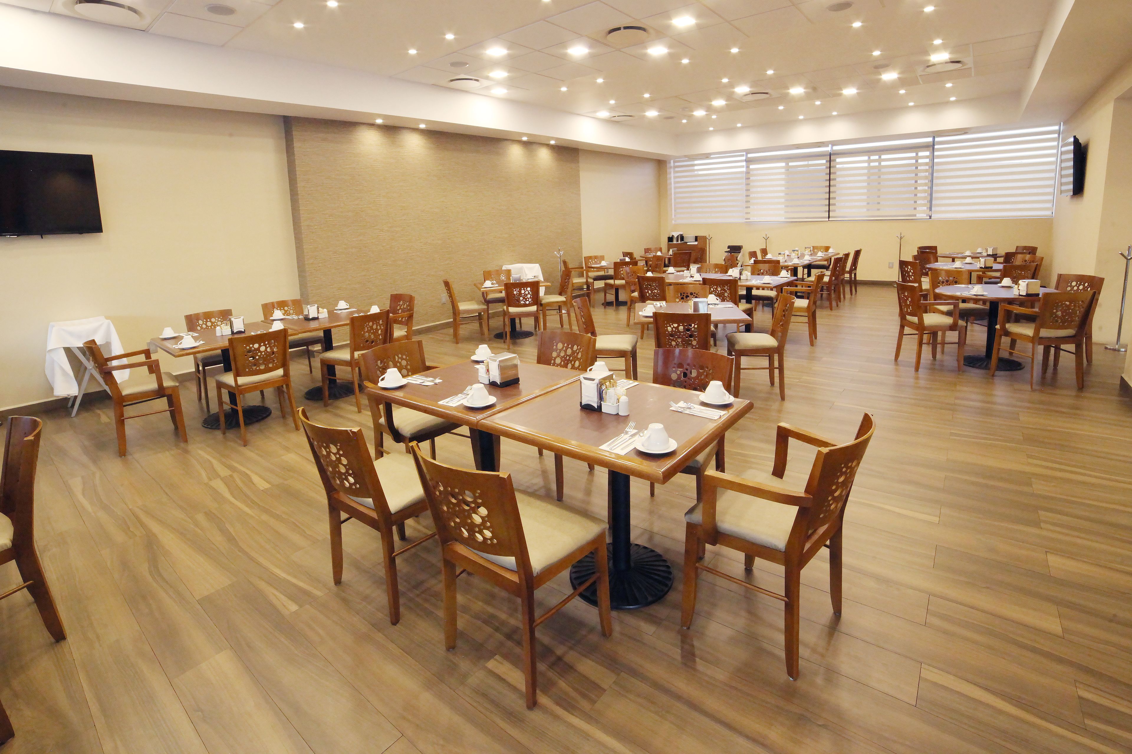 A spacious dining area with multiple tables and chairs, a flat-screen TV mounted on the wall, and large windows with blinds.