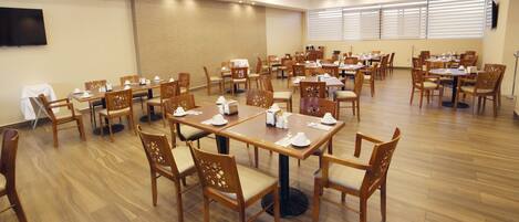 A spacious dining area with multiple tables and chairs, a flat-screen TV mounted on the wall, and large windows with blinds.