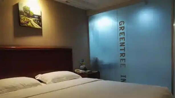 Hotellihuone, jossa on sänky, yöpöytä, taulu seinällä ja seinä, jossa on teksti: ”GREENTREE IN”.
