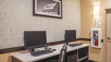 A hotel reception desk with two computer monitors, a keyboard, a mouse, and a chair. There is a framed welcome sign on the wall and a fire extinguisher cabinet.
