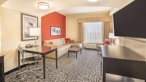 A hotel room with a sofa, a desk, a chair, a television, a window with curtains, and a patterned carpet. - La Quinta Inn & Suites by Wyndham Smyrna TN - Nashville (Smyrna)