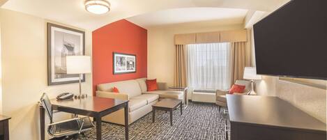 Habitación de hotel con sofá, escritorio, silla, televisor, ventana con cortinas y alfombra estampada.