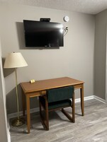 Una habitación con un escritorio de madera, una butaca tapizada en verde, una lámpara de pie y un televisor de pantalla plana montado en la pared.