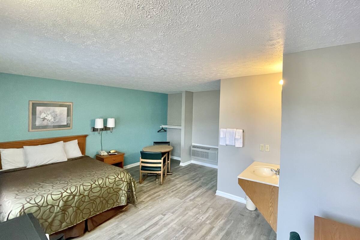 A hotel room with a bed, a desk, a chair, a small table, a lamp, a picture on the wall, and a bathroom with a sink and towel rack.