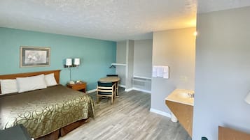 A hotel room with a bed, a desk, a chair, a small table, a lamp, a picture on the wall, and a bathroom with a sink and towel rack.