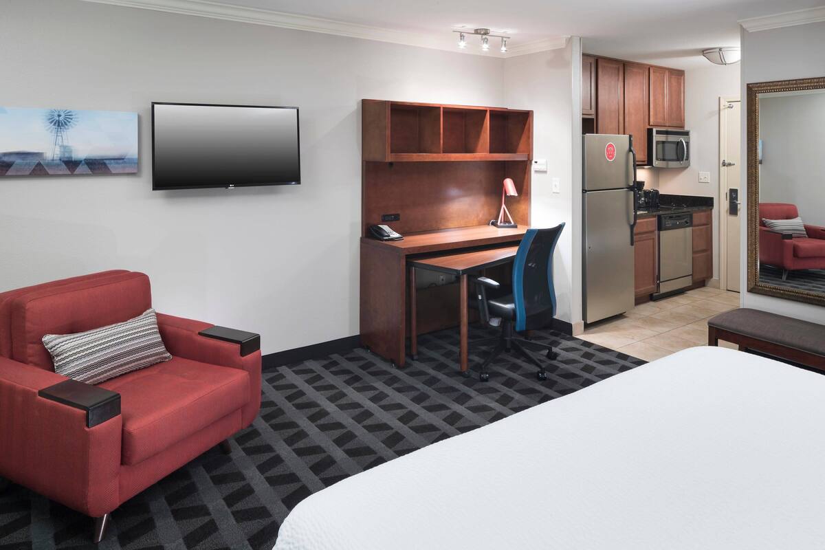 Une chambre d’hôtel comprenant un lit, un fauteuil rouge, un bureau avec une chaise, une télévision, un coin cuisine avec un réfrigérateur, un four à micro-ondes et un four traditionnel, ainsi qu’un tableau accroché au mur.