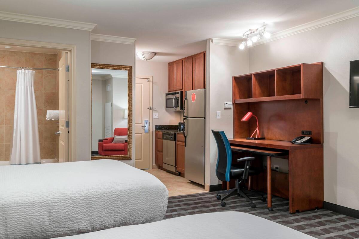 Une chambre d’hôtel avec deux lits, un coin cuisine, un bureau avec une chaise et une lampe rouge.