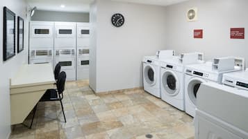 A laundry room with multiple washing machines, a chair, a desk, and a clock on the wall.