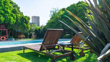 Zona de piscina con dos tumbonas y una mesa, rodeada de exuberante follaje y un edificio al fondo.