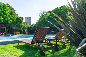 Zona de piscina con dos tumbonas y una mesa, rodeada de exuberante follaje y un edificio al fondo.