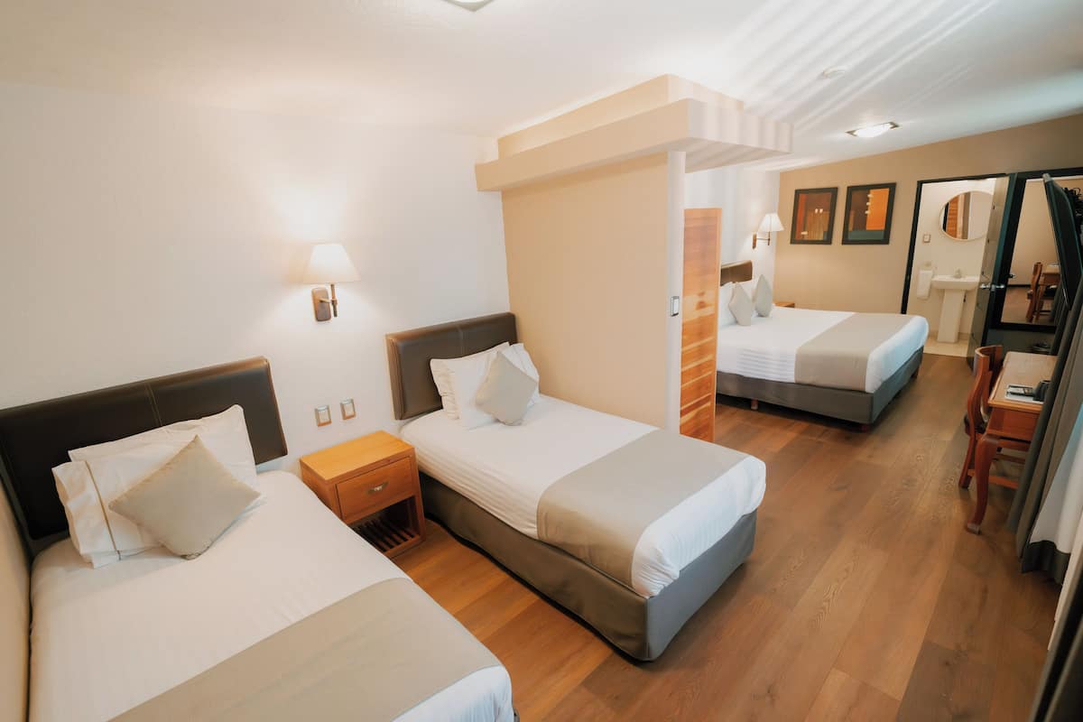 A hotel room with two beds, a wooden floor, a bathroom with a mirror, and a small table with a chair.