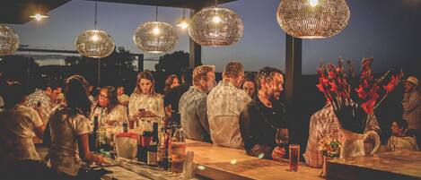 Un bar concurrido con lámparas esféricas colgantes, una barra con diferentes botellas y vasos, y un grupo de personas conversando.