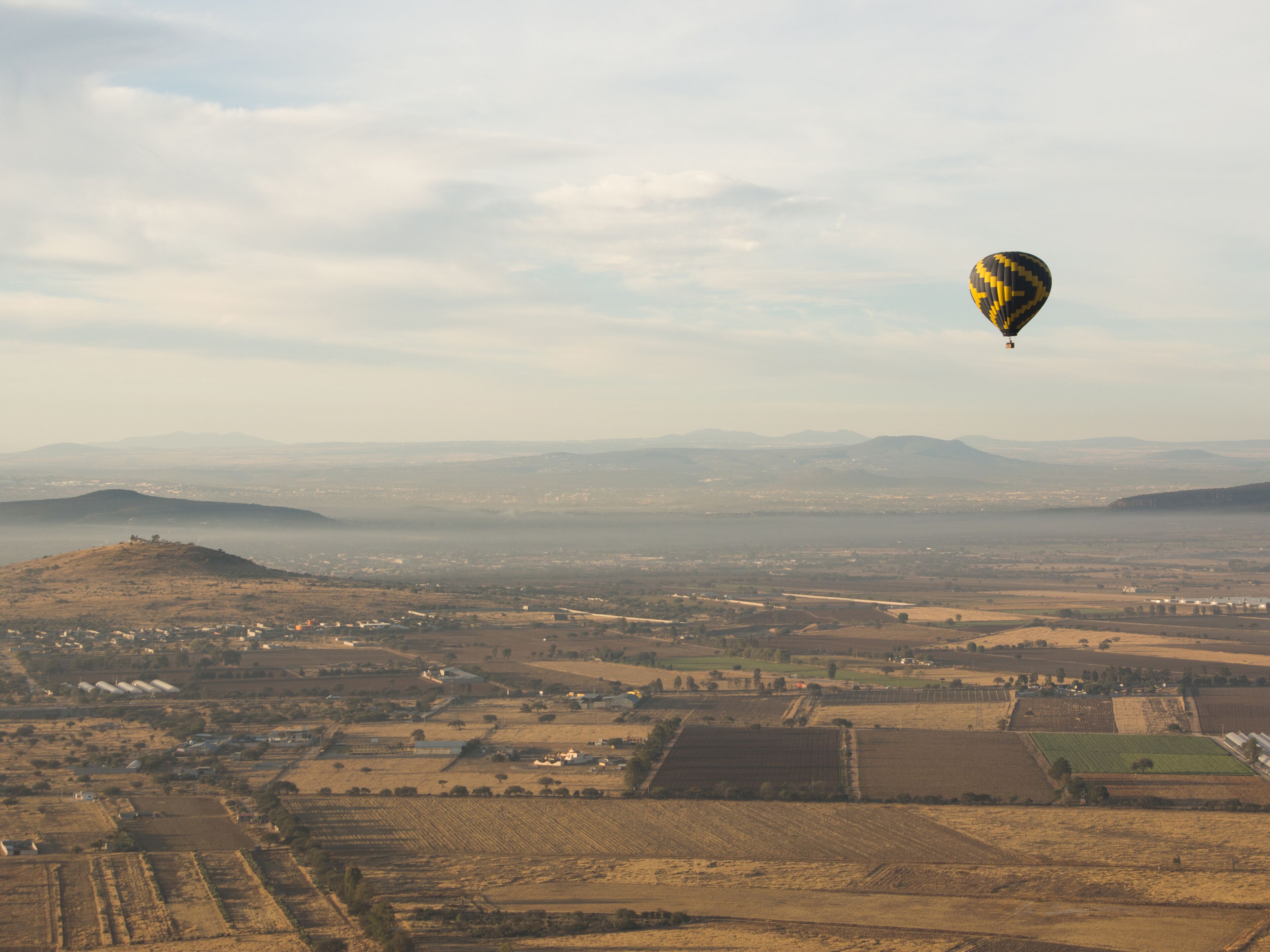 A hot air balloon flying over a rural landscape with fields and distant mountains.