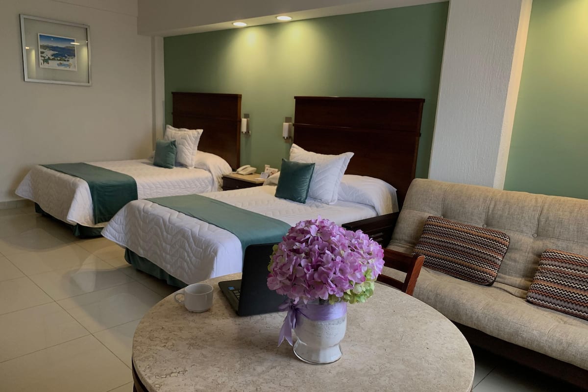 A hotel room with two beds, a sofa, a coffee table with a vase of flowers, and a framed picture on the wall.