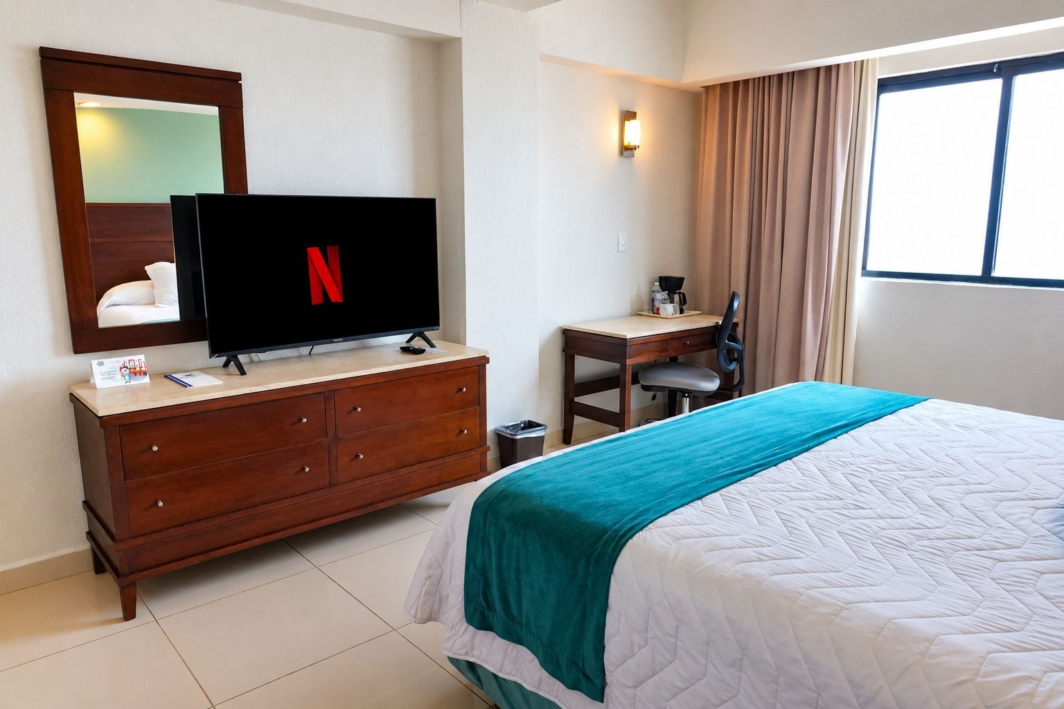 Habitació d'hotel amb llit, escriptori, televisió i finestra amb cortines.