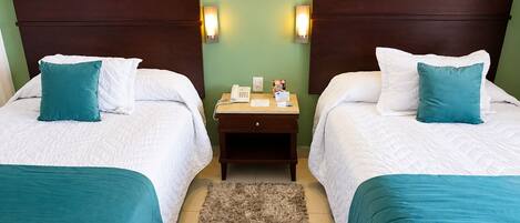 A hotel room with two beds, each with white bedding and teal pillows, a wooden nightstand with a phone and papers, and a fluffy rug on the floor.