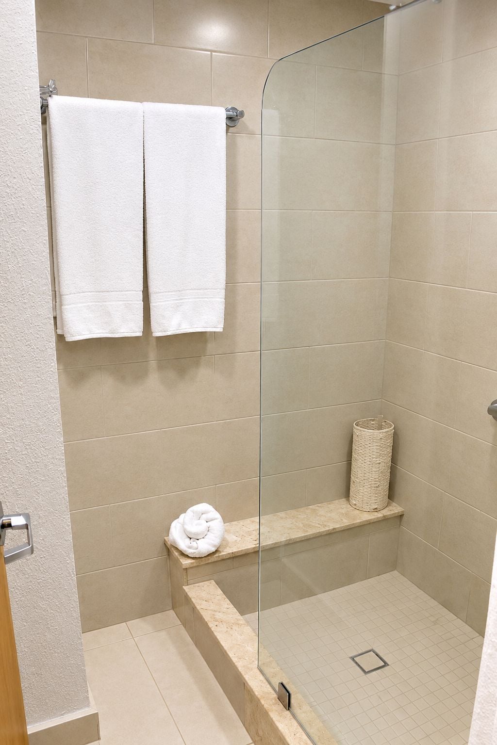 Zona de ducha con azulejos color beige, puerta de cristal, toallero con dos toallas blancas, estante pequeño con una toalla enrollada y una cesta de mimbre.