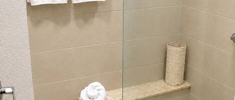 A shower area with beige tiles, a glass door, a towel rack with two white towels, a small shelf with a rolled-up towel, and a wicker basket.