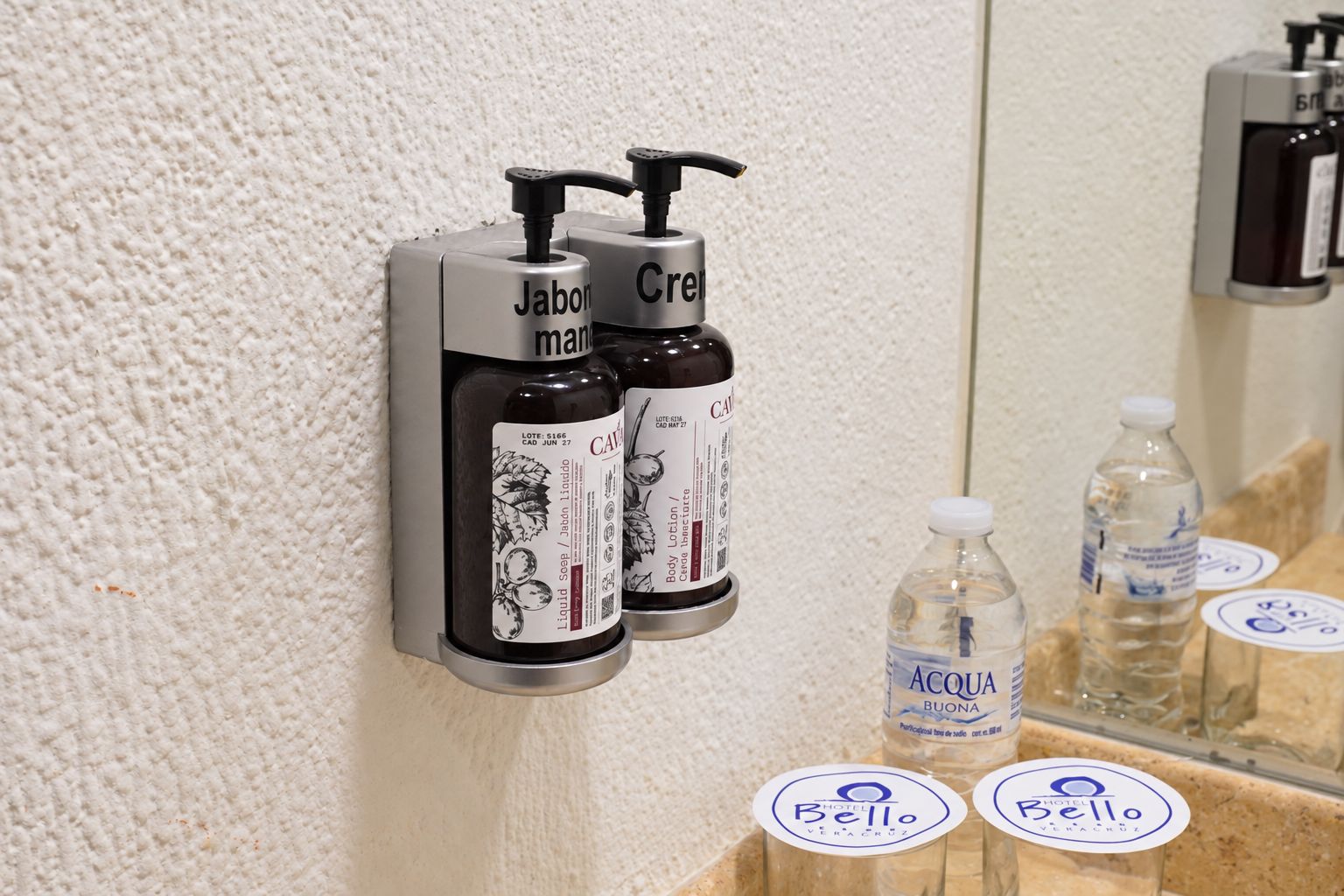 A bathroom with two soap dispensers labeled 'Jabom' and 'Creel' and a mirror reflecting the room.