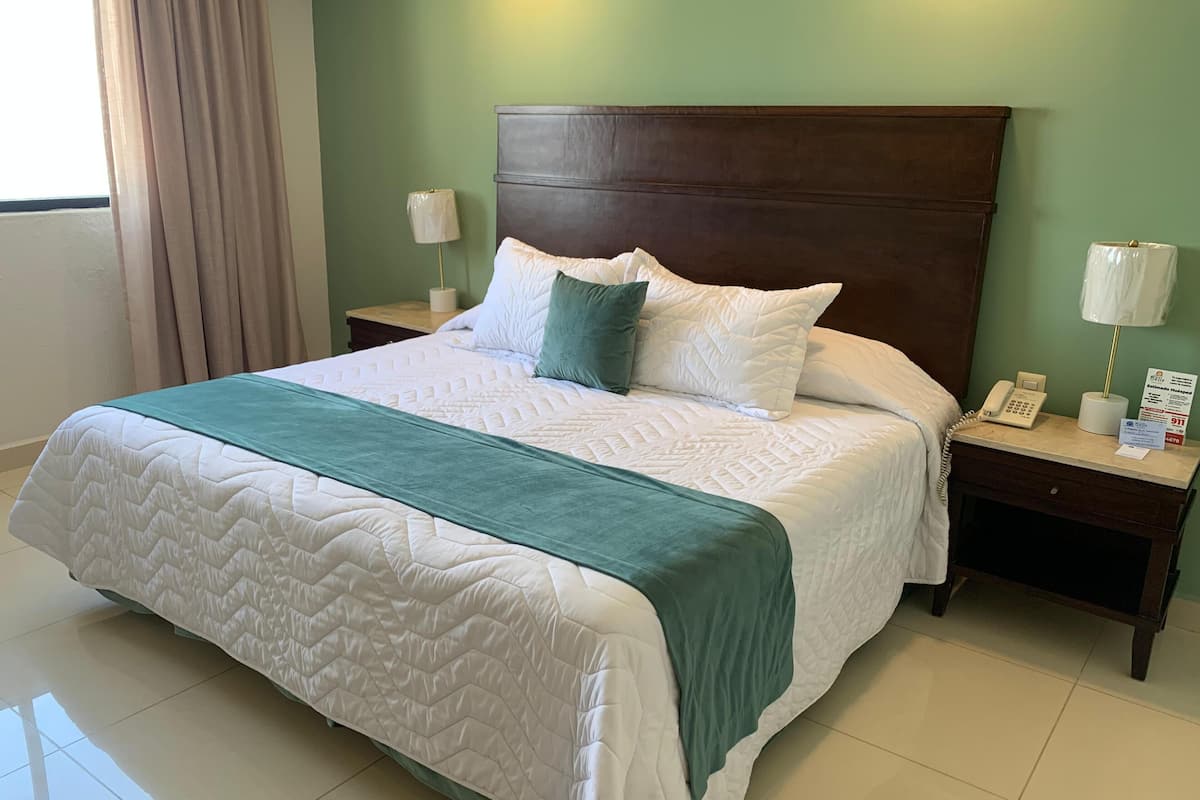 A neatly made bed with a green throw, a wooden headboard, and a nightstand with a lamp and a phone.