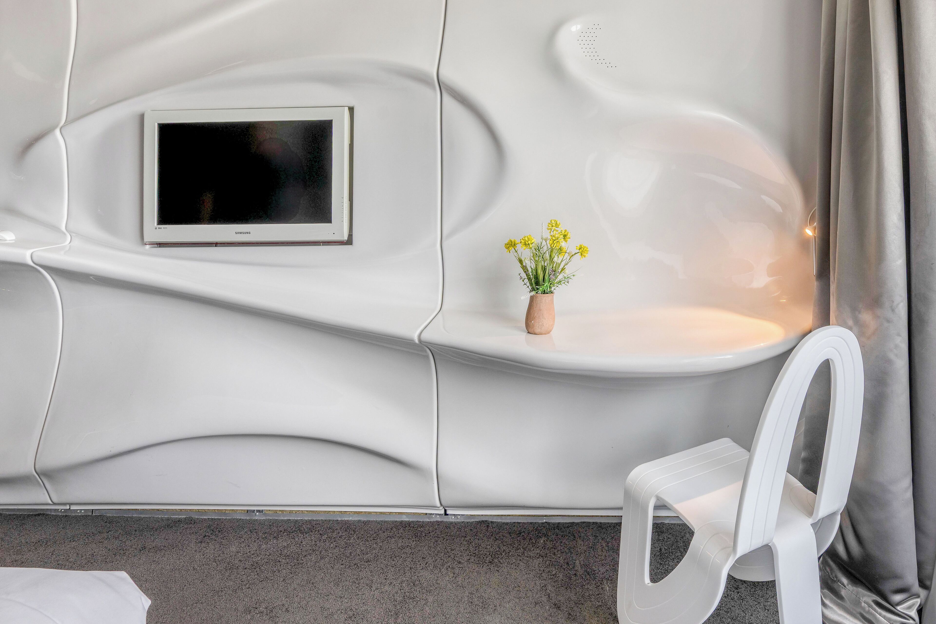 A modern, minimalist room with a curved wall, a flat-screen TV mounted above a small shelf, and a white chair.