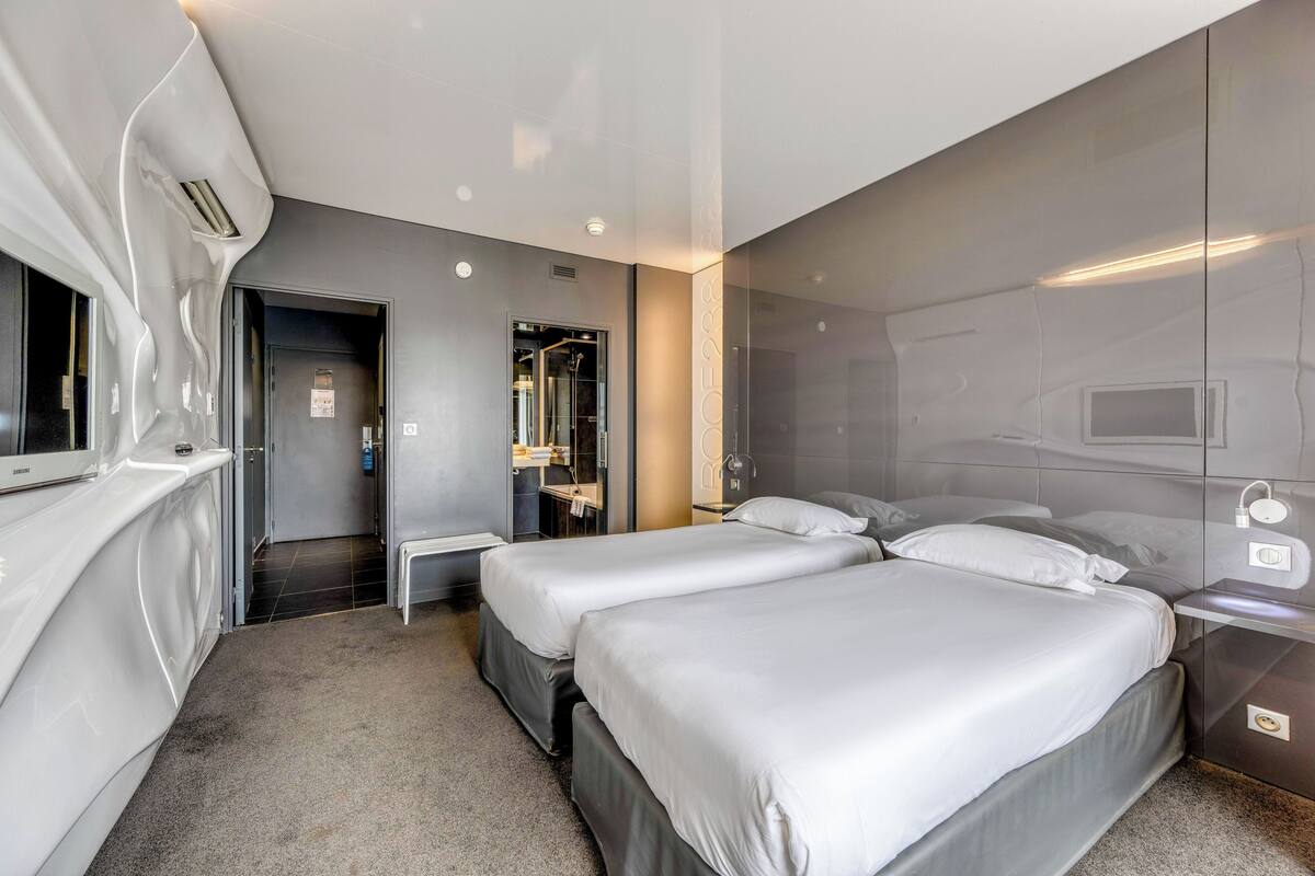 Une chambre d’hôtel moderne avec deux lits, une salle de bain dotée d’un miroir et d’un lavabo, et un petit bureau.