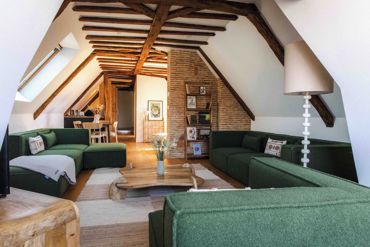 A loft-style living room with green sofas, wooden beams, a sloped ceiling, and a staircase leading to an upper level.