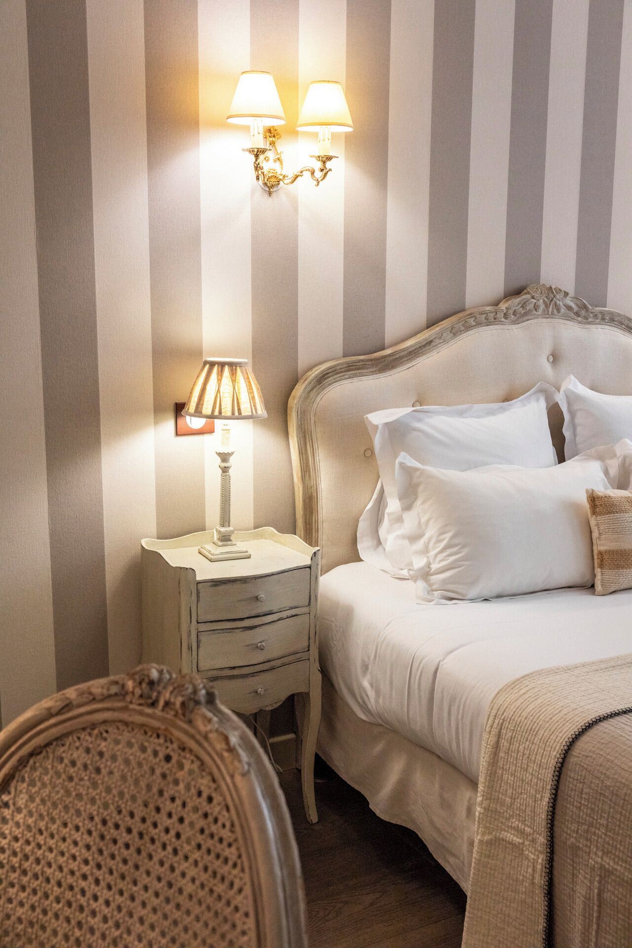 Un dormitorio con papel tapiz a rayas, una cama con ropa blanca, una mesita de noche con una lámpara y una silla de mimbre.
