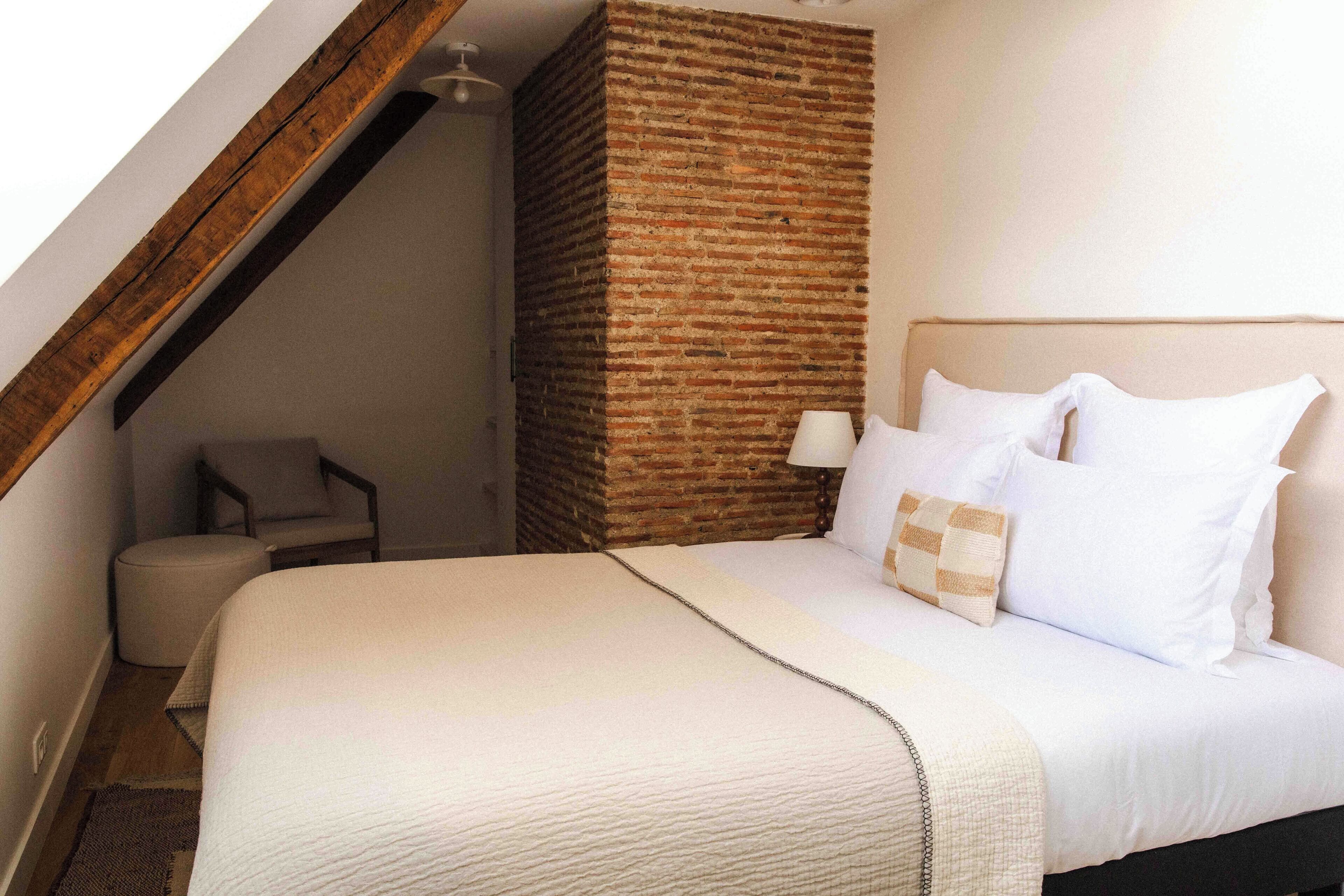 Habitación de hotel con una cama grande, una viga de techo de madera, un muro decorativo de ladrillo y una silla pequeña.