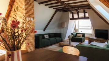 Un salón estilo loft con vigas de madera a la vista, pared de ladrillo y un amplio ventanal. Hay un sofá verde, una mesa de centro de madera y un televisor.