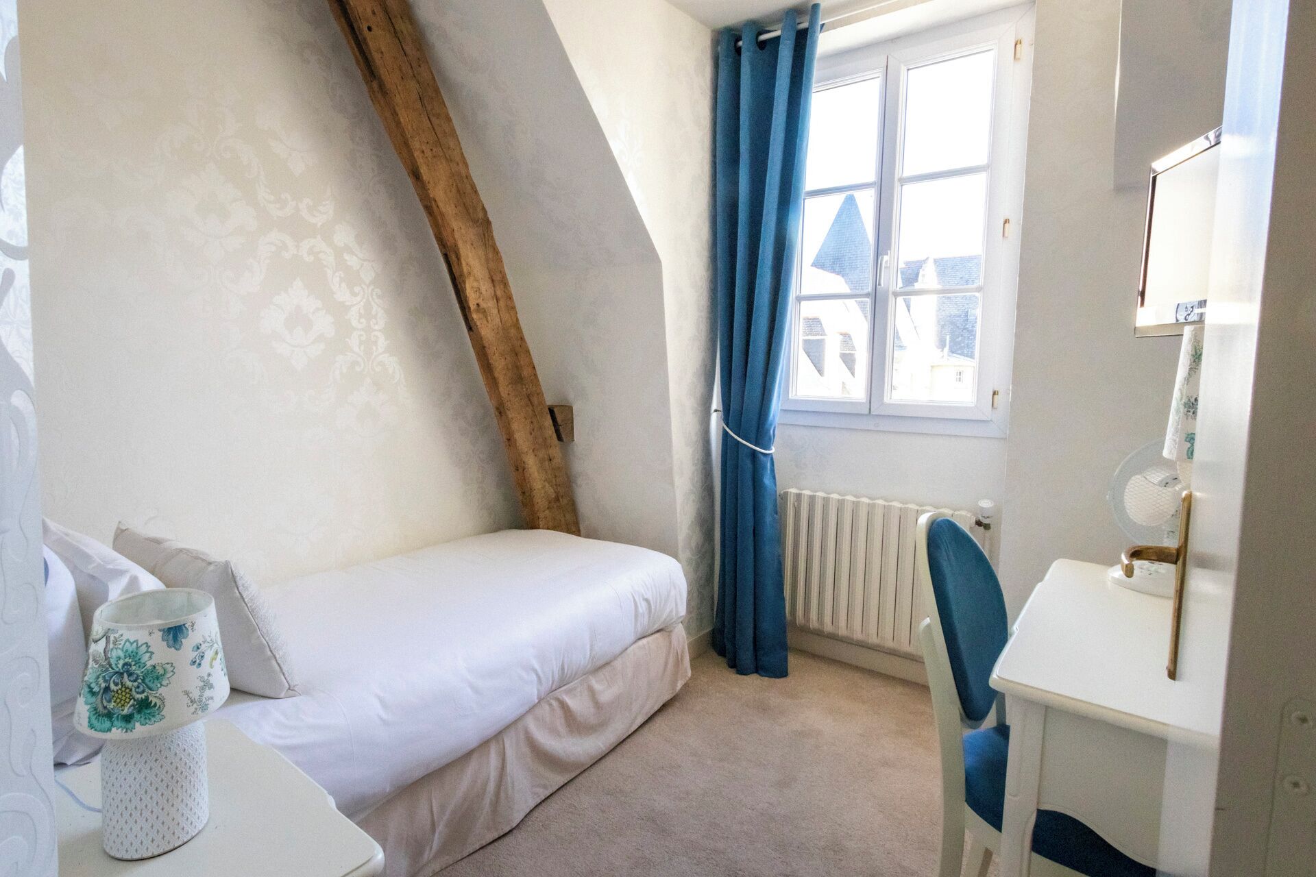 Een kleine, gezellige slaapkamer met een schuine kap, een enkel bed, een bureau met stoel en een raam met blauwe gordijnen.