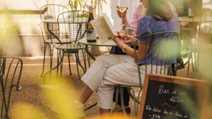 Une personne est assise à une table à l’extérieur, un menu est posé devant elle, et elle est entourée de chaises et d’une plante en pot.