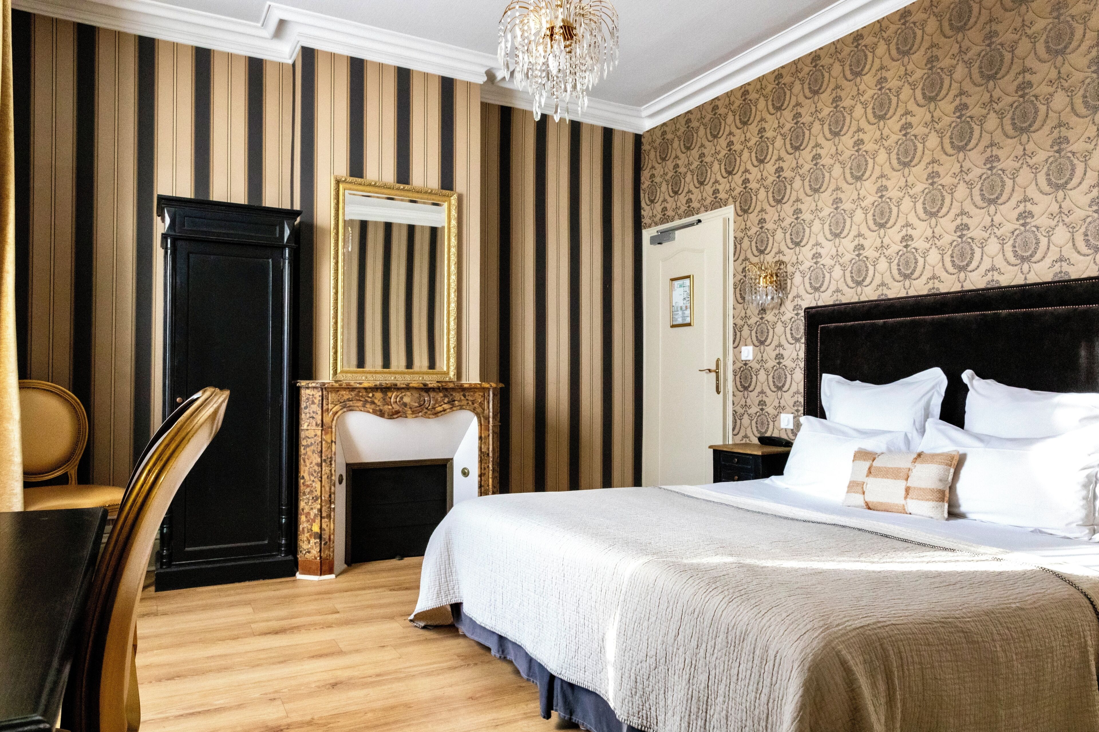 A hotel room with a large bed, a wooden chair, a vanity with a mirror, and a chandelier.