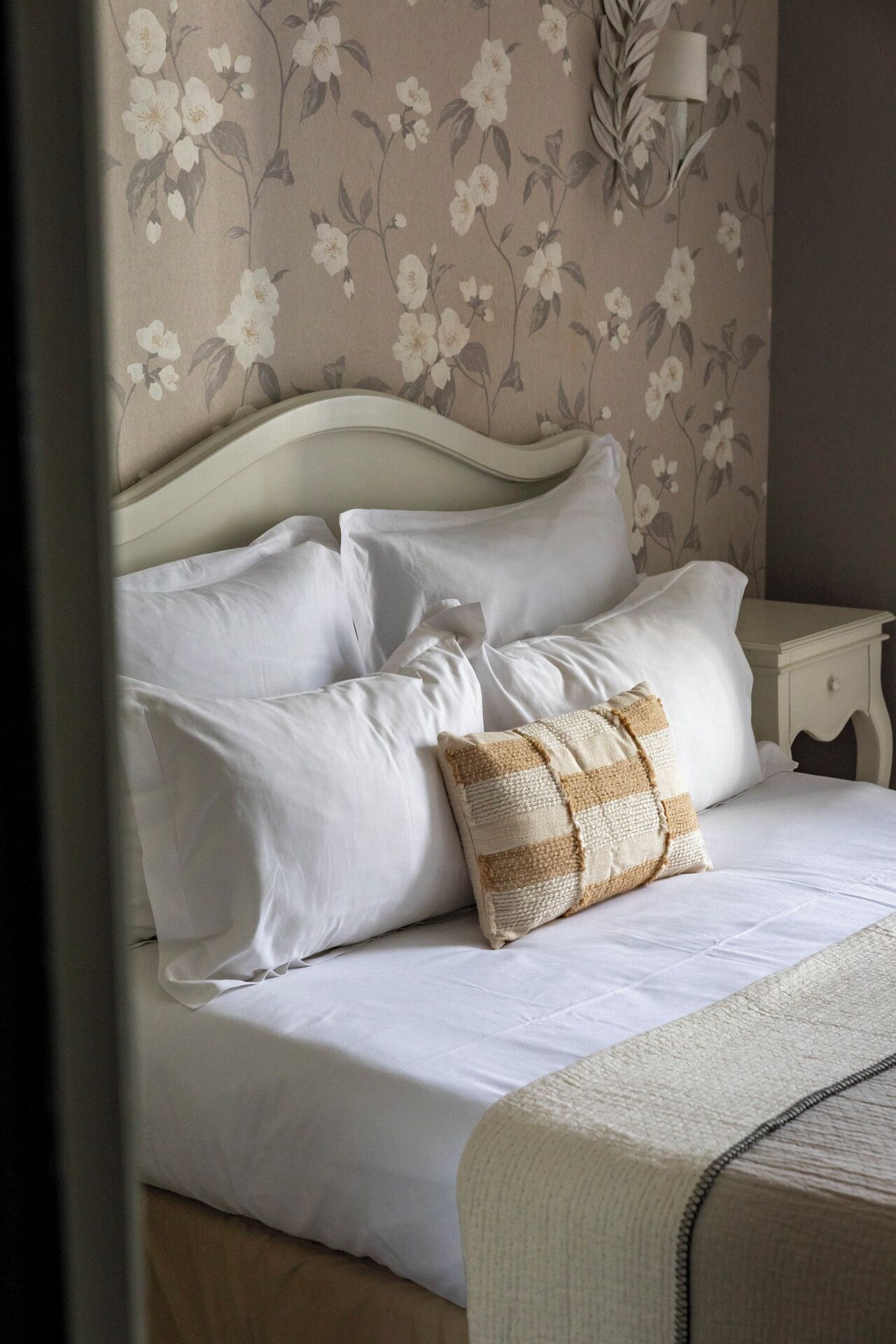 A neatly made bed with white linens and a single patterned pillow, a white nightstand with a lamp, and floral wallpaper.