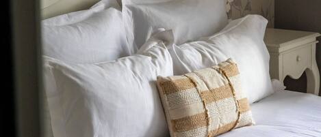 A neatly made bed with white linens and a single patterned pillow, a white nightstand with a lamp, and floral wallpaper.