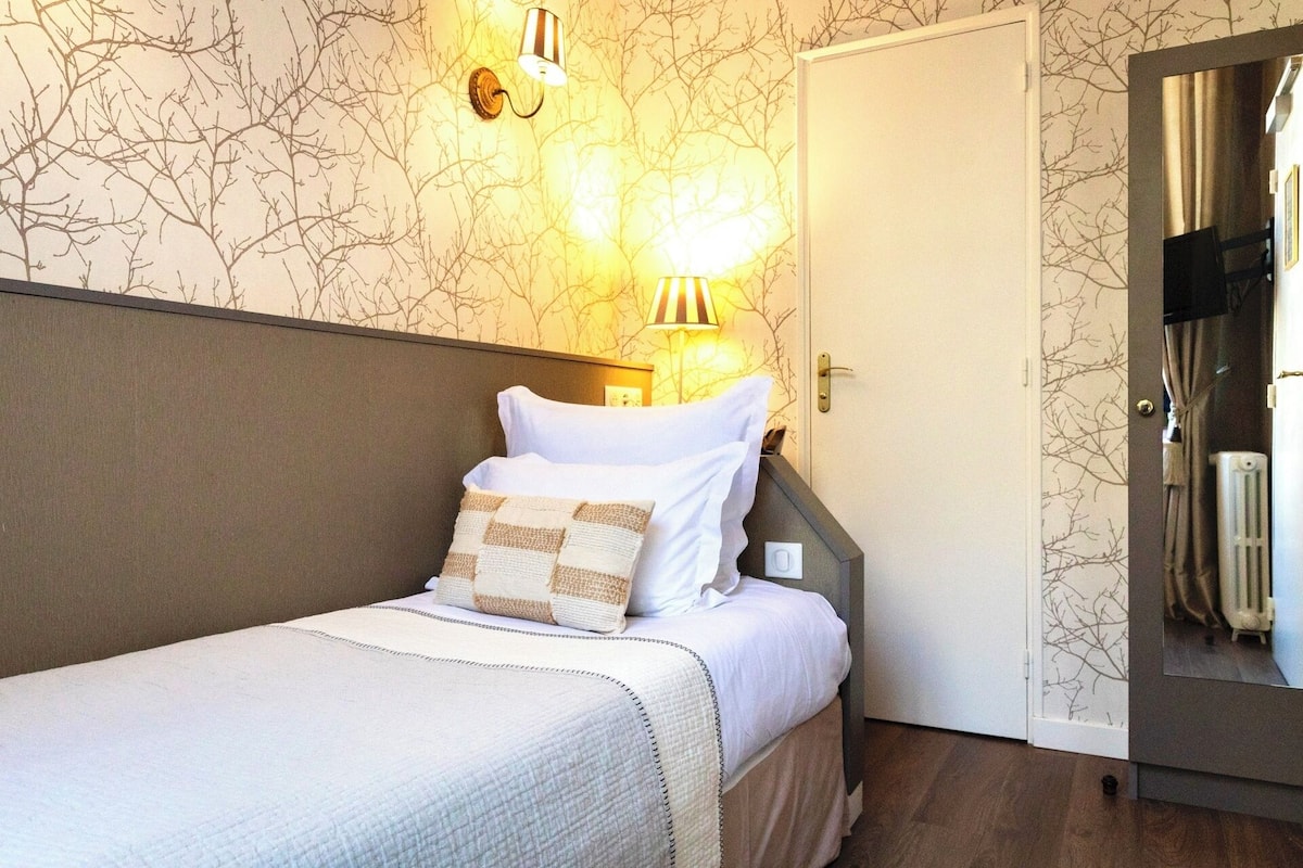 A neatly made bed with white linens and a patterned pillow, a wall-mounted lamp, and a door with a view of a radiator.