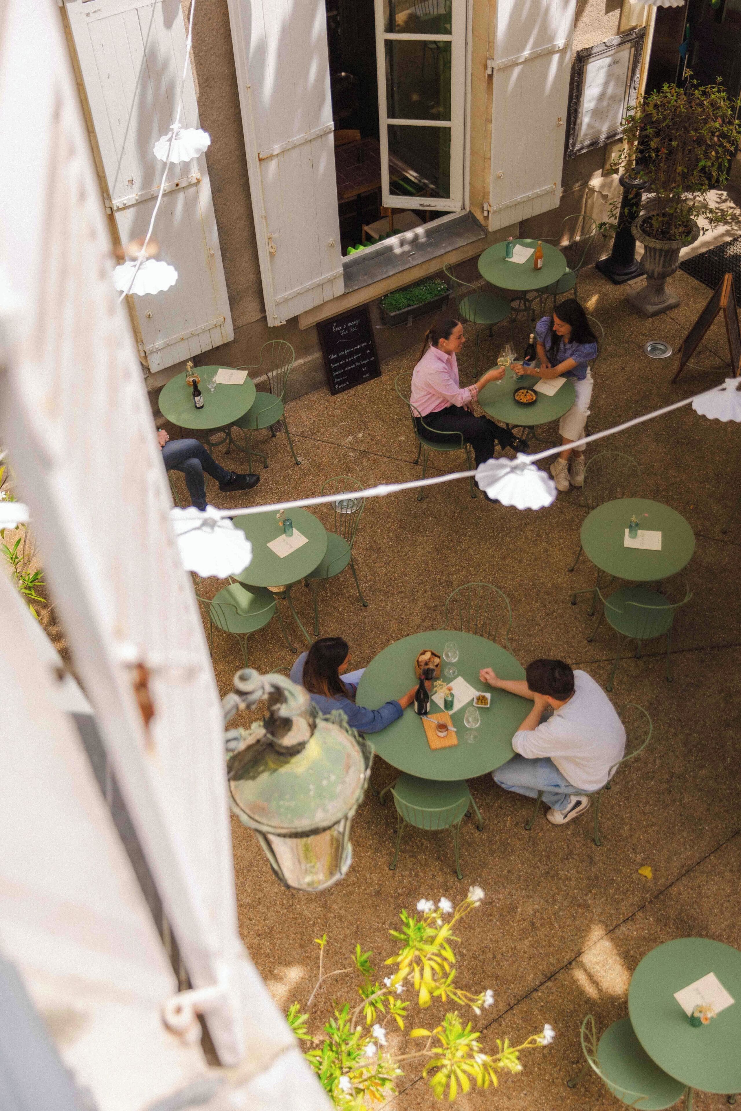 An outdoor patio with multiple green tables and chairs, surrounded by white shutters and a potted plant.