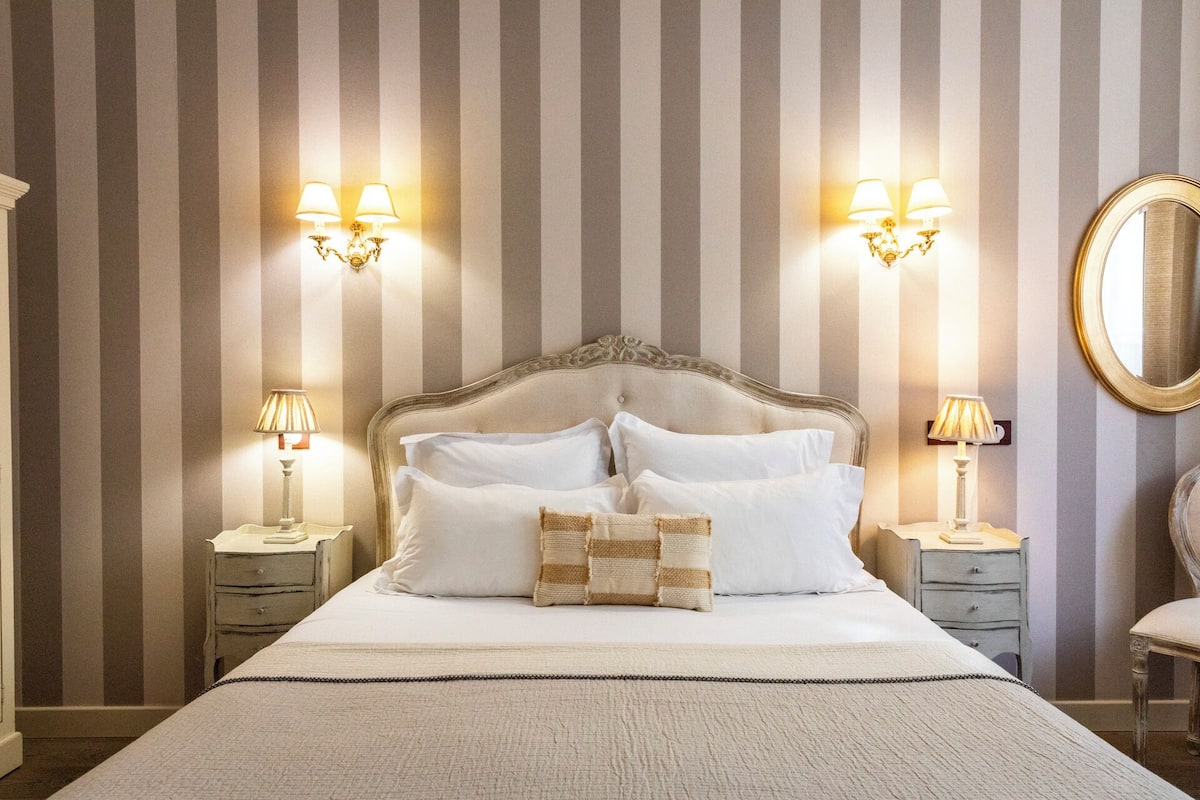 A neatly made bed with white linens, two bedside lamps, and a large mirror on the wall.