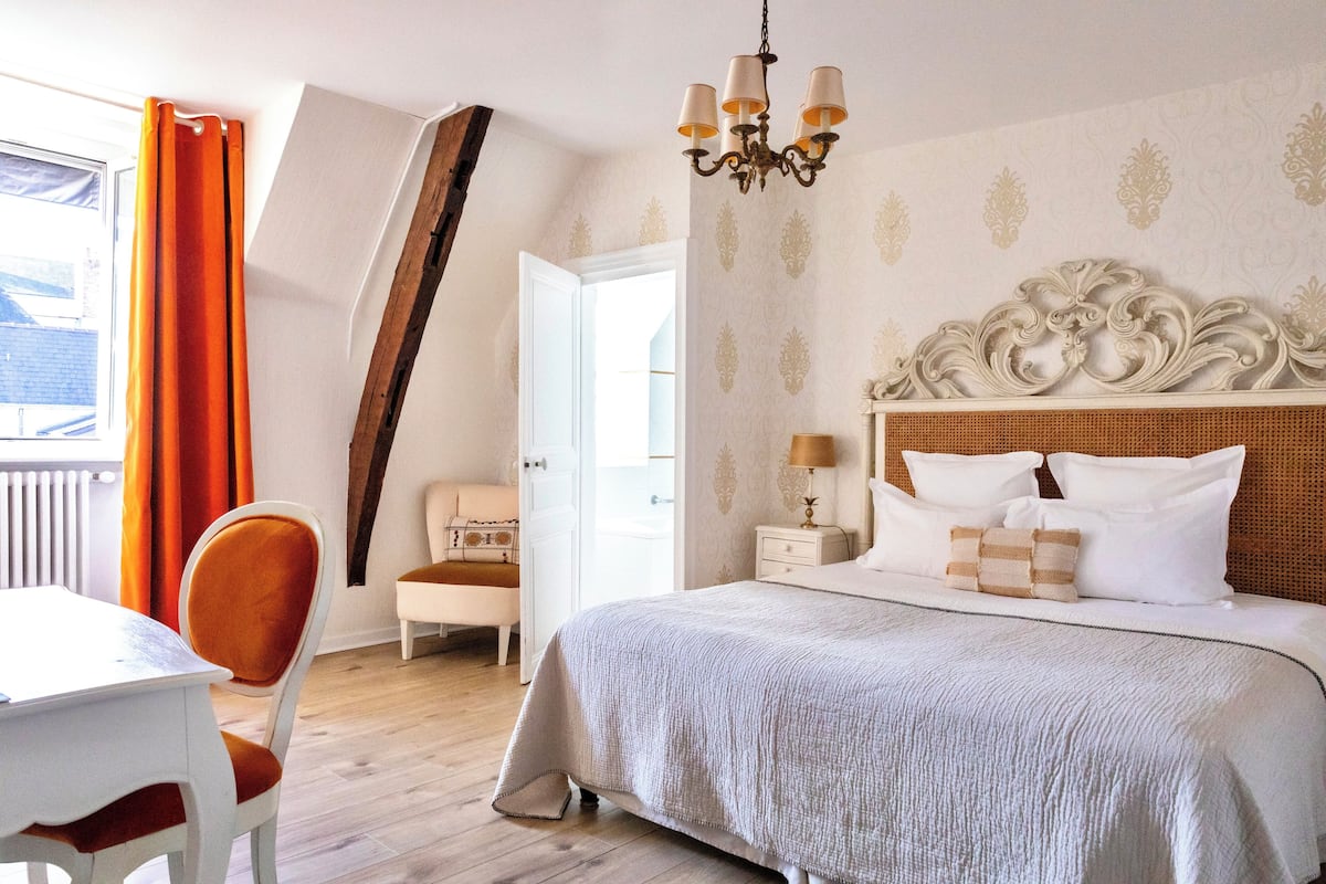 A bedroom with a large bed, a chandelier, a white headboard, and a window with orange curtains.