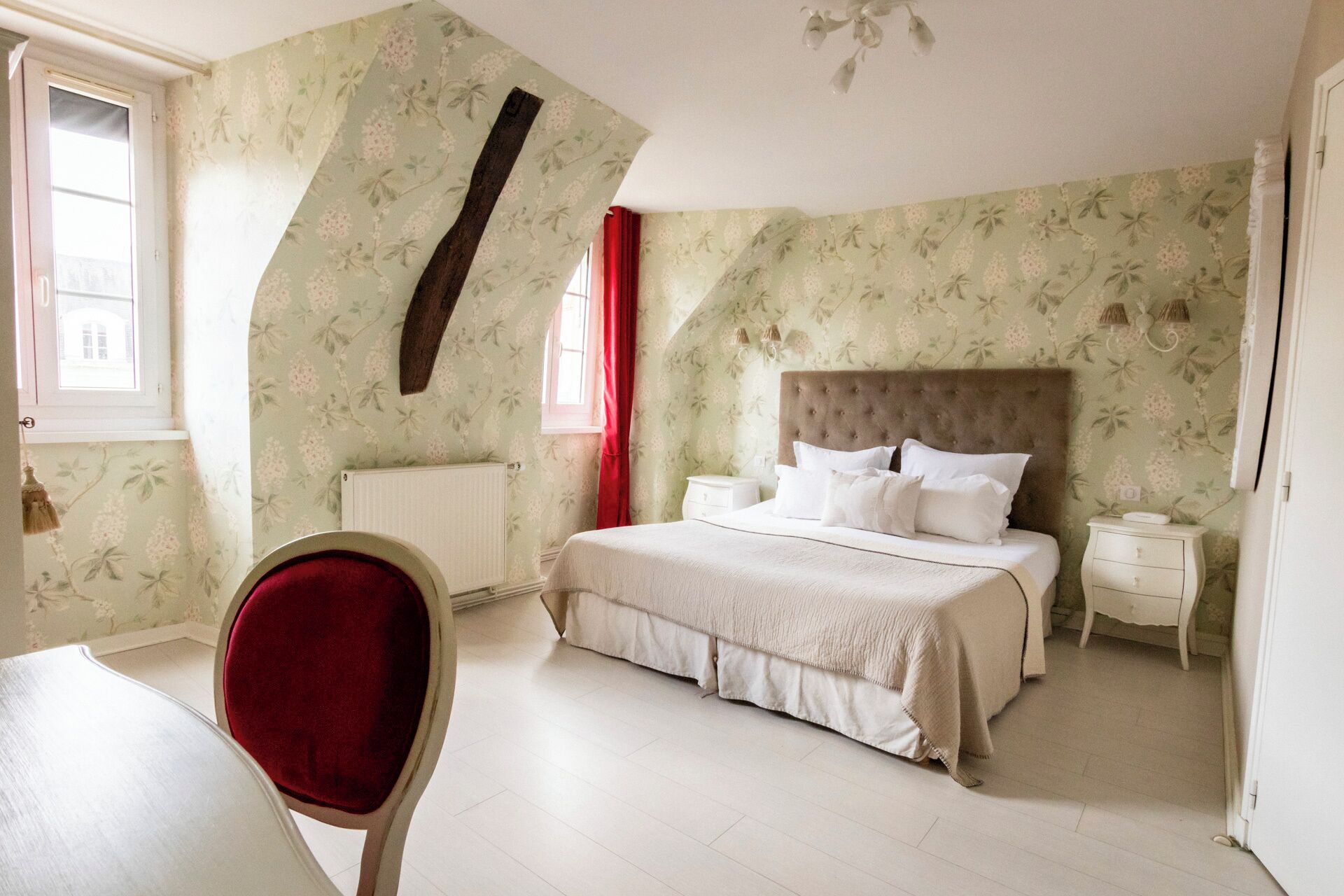 Une chambre à coucher avec un papier peint à motifs floraux, une tête de lit capitonnée, un lit avec du linge de lit blanc, une table de chevet et une chaise rouge.