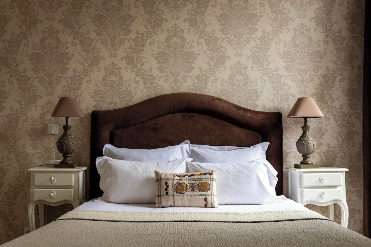 A neatly made bed with white linens, a brown headboard, and decorative pillows. There are two bedside tables with lamps and a wall with a floral pattern.
